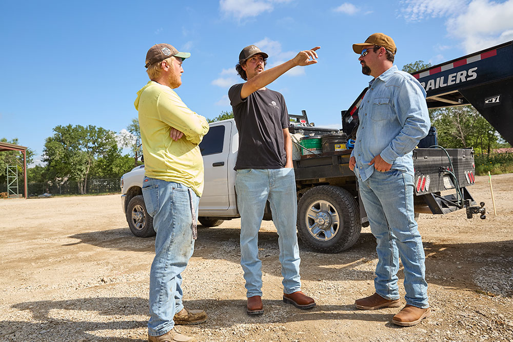 Three men stand talking in a gravel lot near a white pickup truck and a trailer; the man in the center gestures and points into the distance.
